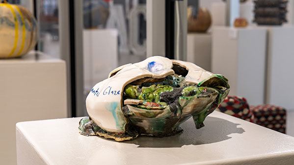 Ceramic shell-like sculpture with a cracked opening revealing green textured interior, displayed on a gallery pedestal.